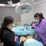 Two dental professionals in scrubs attending to a patient in treatment chair under overhead surgical lighting