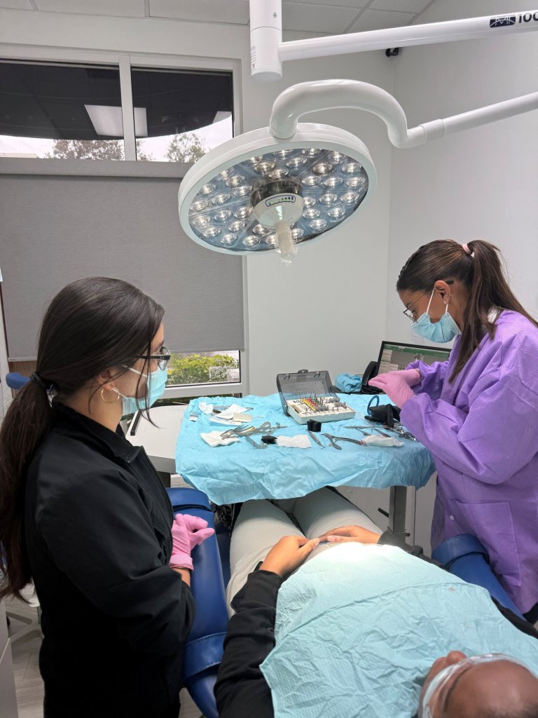 Two dental professionals in scrubs attending to a patient in treatment chair under overhead surgical lighting