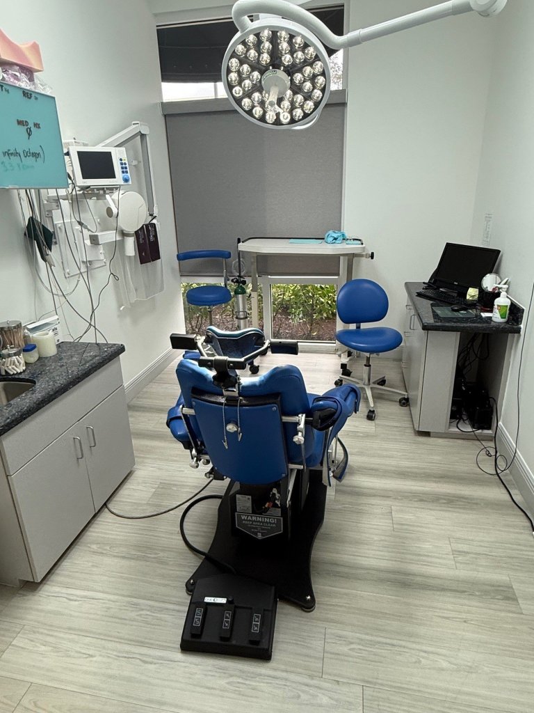 Dental treatment room with blue patient chair, overhead surgical light, cabinetry, and clinical equipment on light wood flooring
