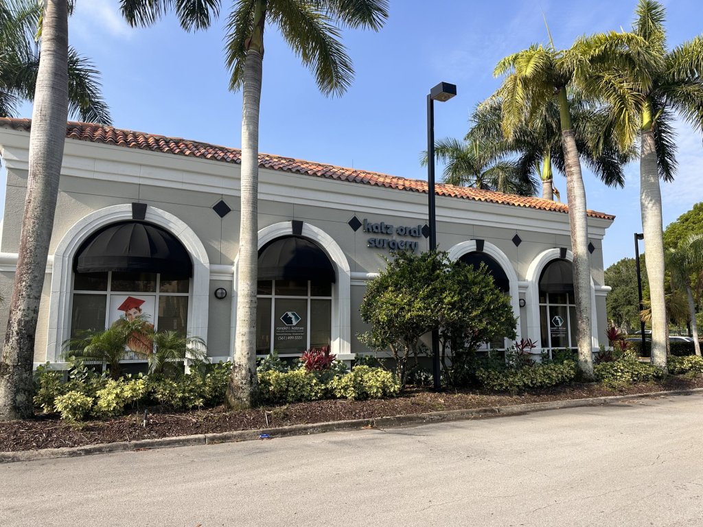 Side angle view of the same professional office building showing white arched colonnade, tropical landscaping with palm trees and shrubs, and paved parking area under blue sky