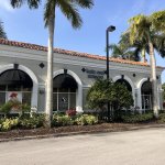 Side angle view of the same professional office building showing white arched colonnade, tropical landscaping with palm trees and shrubs, and paved parking area under blue sky