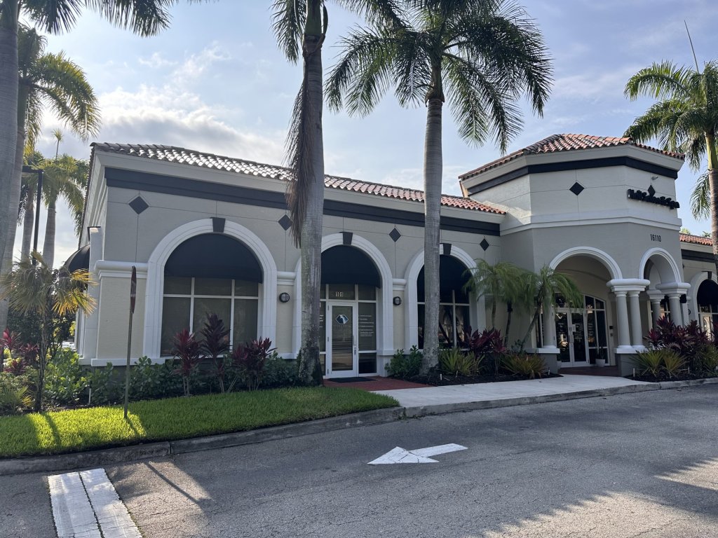 Exterior view of Mediterranean-style medical/dental office building with white stucco walls, terracotta roof tiles, arched windows and entryways, surrounded by palm trees and professional landscaping